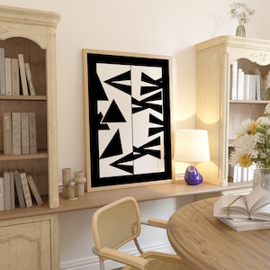 May include: A framed abstract art print featuring a black and white geometric design of triangles and other shapes. The artwork is displayed in a light-colored wooden frame. The print is in a room with bookshelves, a lamp, and a round table.