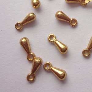 May include: A group of gold-colored teardrop-shaped metal charms with a loop at the top. The charms are scattered on a white surface.
