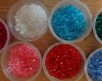 May include: Seven small containers filled with colorful, faceted glass beads. The containers are arranged in a circle on a wooden surface. The beads are clear, red, pink, blue, green, and turquoise.