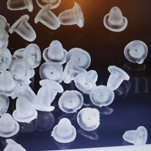 Peut inclure: Une image en gros plan d'une grande quantité de dos d'oreille en plastique transparent. Les dos sont en forme de petits dômes plats.