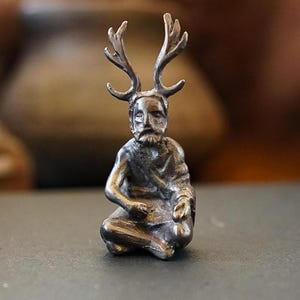 以下が含まれることがあります： 頭の上に角のある人の小さな銀色の置物。その人は足を組んで座り、手に何かを持っています。