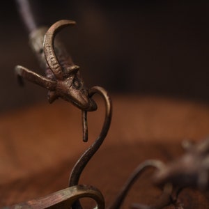 Peut inclure: Gros plan d'une sculpture en bronze d'une chèvre avec la tête penchée vers le bas, comme si elle buvait dans une base métallique incurvée. La sculpture est posée sur une surface en bois.