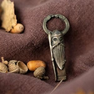 May include: A silver-toned metal Viking-themed bottle opener with a detailed design of a warrior with a beard and helmet. The opener is lying on a brown fabric surface with acorns scattered around it.