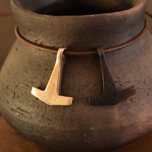 Peut inclure: Deux colliers avec des pendentifs en forme de marteau. L'un est doré, l'autre gris foncé. Les deux sont suspendus à un cordon en cuir marron. Les colliers sont présentés sur un vase en céramique gris foncé texturé.