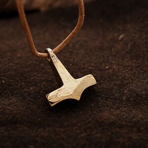 Könnte beinhalten: Ein silberfarbener Anhänger in Form von Thors Hammer, an einer braunen Lederschnur befestigt. Der Hammer hat ein detailliertes Design und wird vor einem dunkelbraunen Hintergrund präsentiert, was ein rustikales oder Wikinger-Thema suggeriert.