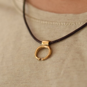 Può includere: Una semplice collana con un ciondolo dorato a forma di piccolo cerchio aperto. La collana è su un cordoncino nero.
