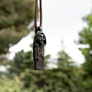 May include: A black metal pendant depicting a hooded figure with a skeletal face, hanging from a brown leather cord. The pendant is in focus, while the background is blurred.