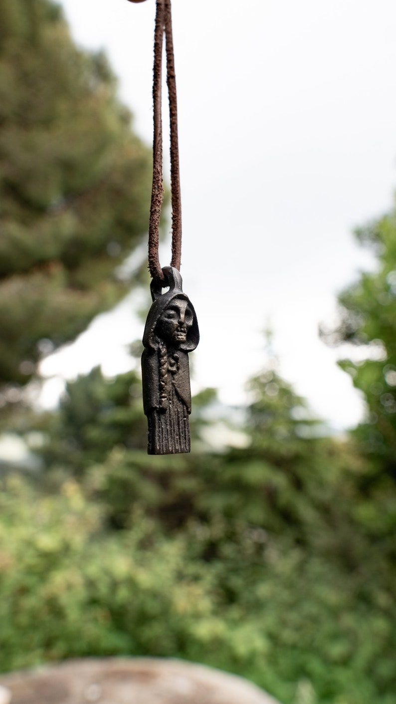 May include: A black metal pendant depicting a hooded figure with a skeletal face, hanging from a brown leather cord. The pendant is in focus, while the background is blurred.