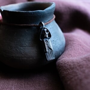 May include: A dark gray ceramic pot with a brown leather cord and a silver skull pendant. The pot is sitting on a brown fabric surface.