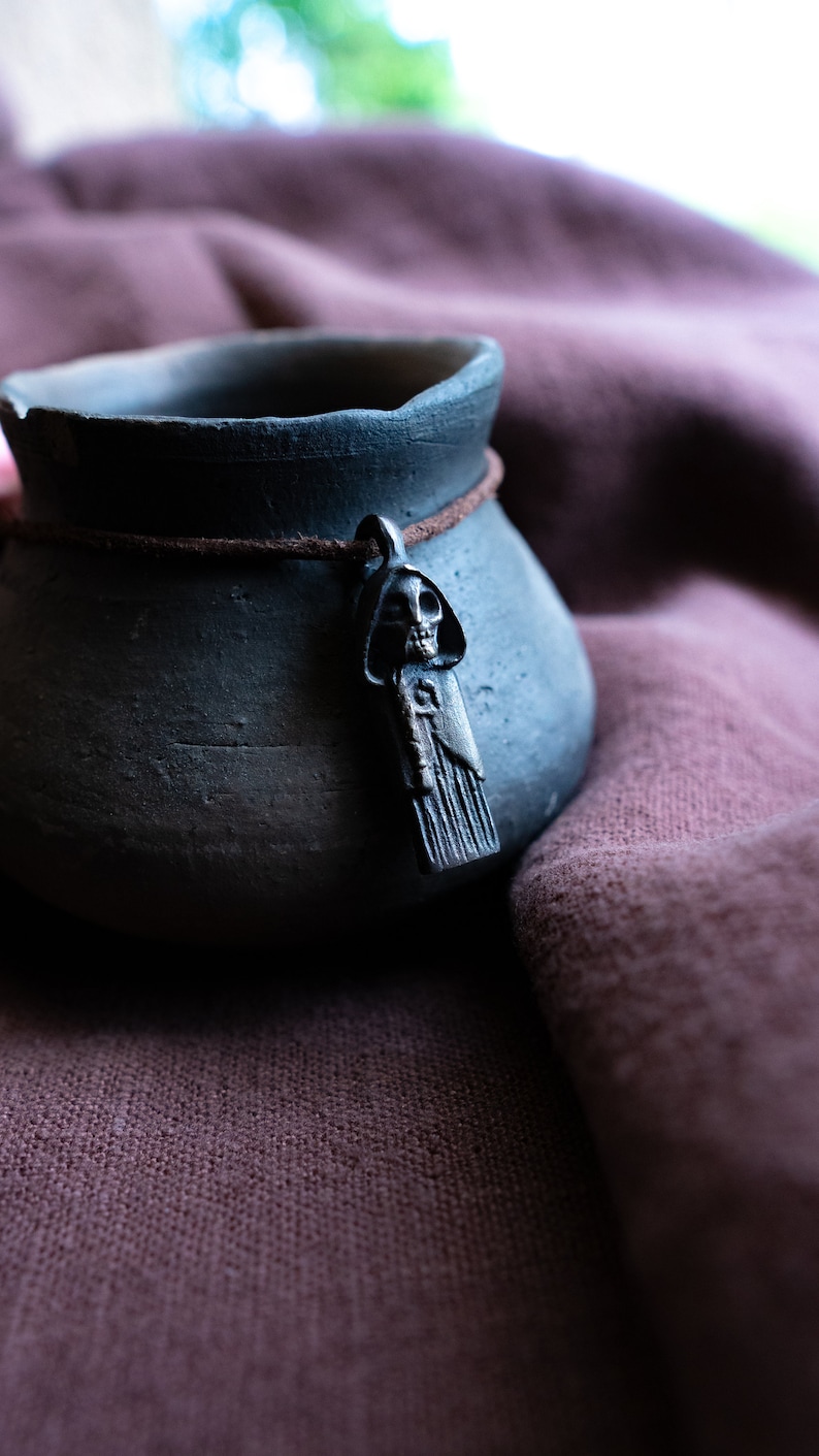 May include: A dark gray ceramic pot with a brown leather cord and a silver skull pendant. The pot is sitting on a brown fabric surface.