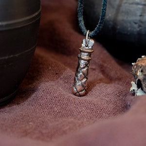 Può includere: Collana con pendente color argento dal design cilindrico e strutturato. Il pendente è appeso a un cordino nero. Lo sfondo comprende tessuto marrone scuro, un contenitore nero e un pezzo di legno.