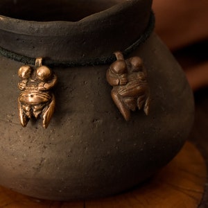 Può includere: Due ciondoli in bronzo con un design a figura umana pendono da un cordino in pelle marrone scuro attorno a un vaso in ceramica marrone scuro. Il vaso è appoggiato su una superficie di legno.