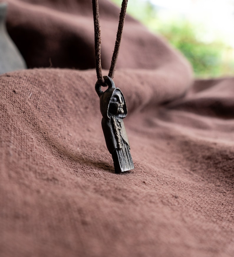 May include: A black metal pendant necklace featuring a hooded figure with a skeletal face. The pendant is hanging on a brown leather cord.