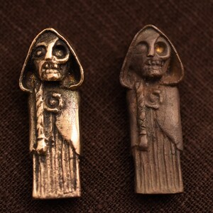 May include: Two small, detailed, bronze sculptures of hooded figures with skeletal faces. The figure on the left has a black eye, while the figure on the right has a gold eye.