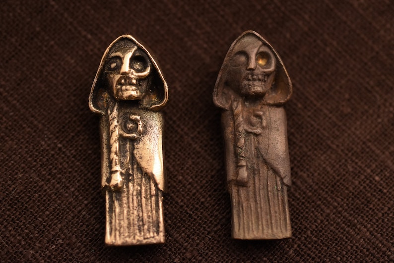 May include: Two small, detailed, bronze sculptures of hooded figures with skeletal faces. The figure on the left has a black eye, while the figure on the right has a gold eye.