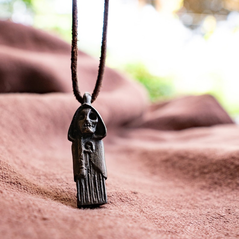 May include: A dark brown leather cord necklace with a detailed metal pendant depicting a hooded figure with a skull face.