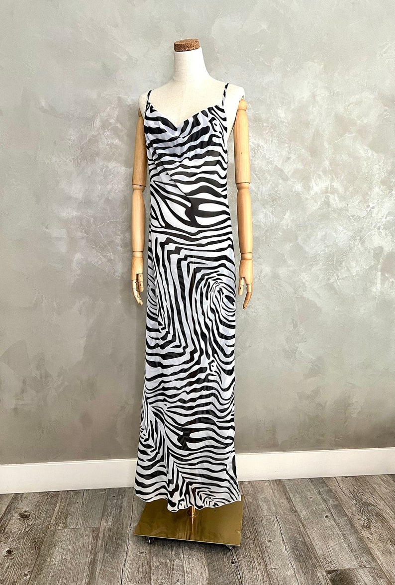 May include: A floor-length dress with a zebra print in black and white. The dress has thin spaghetti straps and a cowl neckline. The dress is displayed on a mannequin with wooden arms and a cork head.