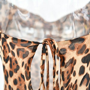 May include: A close-up of a leopard print dress. The dress features thin spaghetti straps and a tie detail at the back. The fabric is a sheer material with a brown and black spotted pattern.