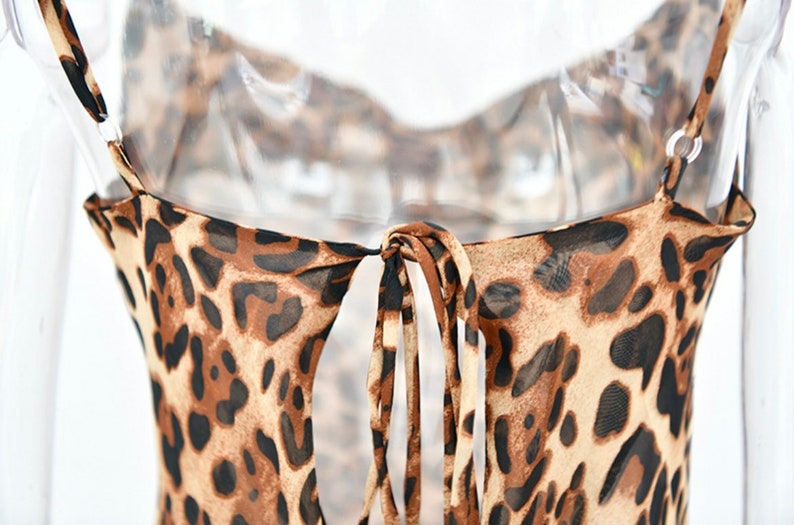 May include: A close-up of a leopard print dress. The dress features thin spaghetti straps and a tie detail at the back. The fabric is a sheer material with a brown and black spotted pattern.