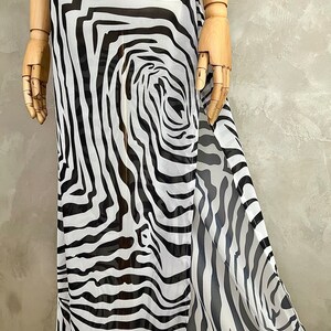 May include: A flowing, floor-length dress with a bold zebra print in black and white. The fabric appears lightweight and semi-sheer, with a split detail on one side. The dress is displayed on a wooden mannequin.
