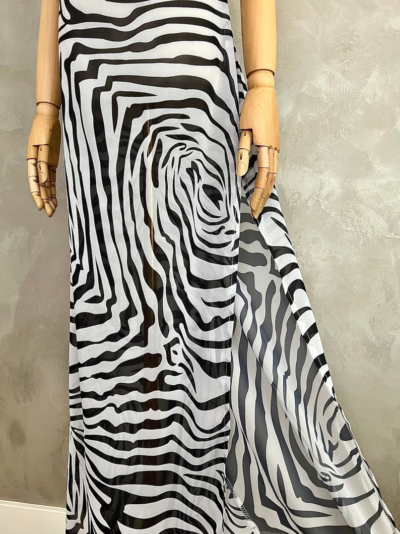 May include: A flowing, floor-length dress with a bold zebra print in black and white. The fabric appears lightweight and semi-sheer, with a split detail on one side. The dress is displayed on a wooden mannequin.