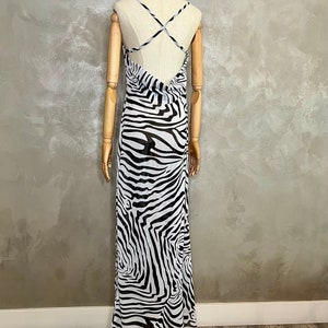 May include: A long, flowing dress with a zebra print in black and white. The dress features a low back and thin straps that cross in the back. The dress is displayed on a mannequin.
