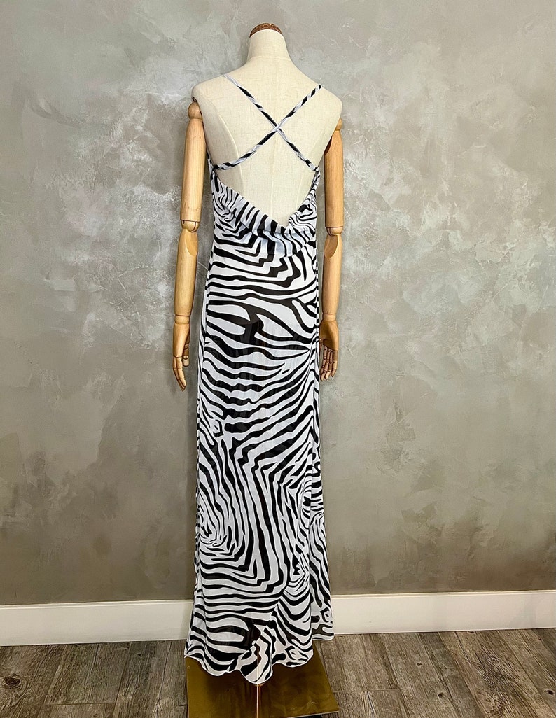 May include: A long, flowing dress with a zebra print in black and white. The dress features a low back and thin straps that cross in the back. The dress is displayed on a mannequin.