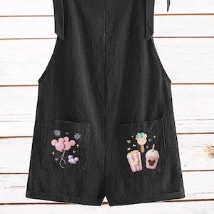 Op de afbeelding: Zwarte romper met strikbandjes en geborduurde zakdetails. De zakken hebben kleurrijke geborduurde ontwerpen van ballonnen, ijs en popcorn. De romper is gemaakt van een zachte, lichtgewicht stof.