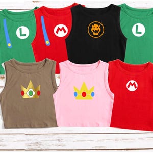 May include: A collection of sleeveless shirts in various colors, each featuring a different character design. The shirts include green with a white "L", red with a white "M", black with a Bowser symbol, and more. The shirts are displayed on a light wood surface.