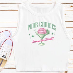 May include: White sleeveless crop top with a graphic design. The design features the text "Pour Choices" above a cocktail glass with a pink sphere and lime slice, with the text "Around the World!" below. A pair of white sneakers are in the lower left corner.