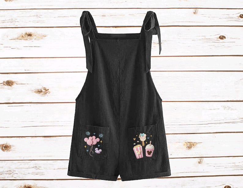 Op de afbeelding: Zwarte romper met strikbandjes en geborduurde zakdetails. De zakken hebben kleurrijke geborduurde ontwerpen van ballonnen, ijs en popcorn. De romper is gemaakt van een zachte, lichtgewicht stof.