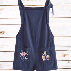 Op de afbeelding: Marineblauwe romper met strikbandjes en geborduurde zakdetails. E&eacute;n zak heeft roze ballonnen en de andere een popcorn-, drank- en Mickey Mouse-ontwerp. De jumpsuit in shorts-stijl is gemaakt van een zachte stof.