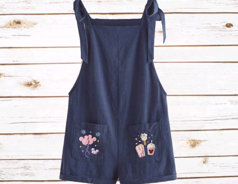 Op de afbeelding: Marineblauwe romper met strikbandjes en geborduurde zakdetails. E&eacute;n zak heeft roze ballonnen en de andere een popcorn-, drank- en Mickey Mouse-ontwerp. De jumpsuit in shorts-stijl is gemaakt van een zachte stof.