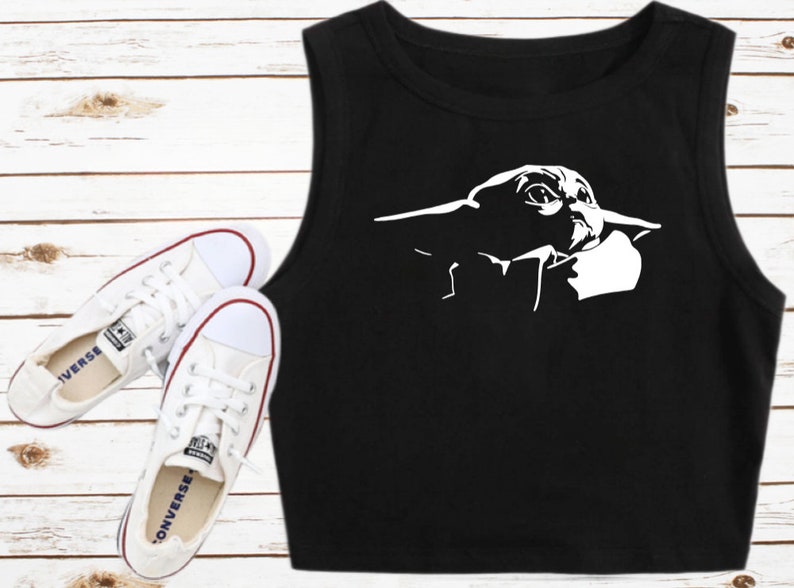 May include: Black crop top featuring a white graphic of a character from a popular science fiction franchise. The top is sleeveless and has a round neckline. White canvas sneakers with red accents are also in the image.