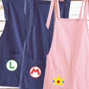 May include: Three sleeveless rompers with tie-shoulder straps and two front pockets. Two rompers are dark blue, one featuring a green letter L and the other a red letter M. The third romper is light pink with a yellow crown graphic on the pocket.