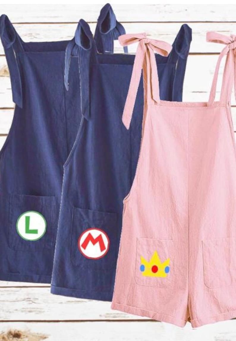 May include: Three sleeveless rompers with tie-shoulder straps and two front pockets. Two rompers are dark blue, one featuring a green letter L and the other a red letter M. The third romper is light pink with a yellow crown graphic on the pocket.