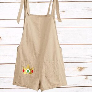 May include: A beige overall romper featuring tie-shoulder straps and two front pockets. The left pocket displays a graphic of a gold crown with a green center gem and red side accents, providing a distinct visual detail on the solid light-colored fabric.