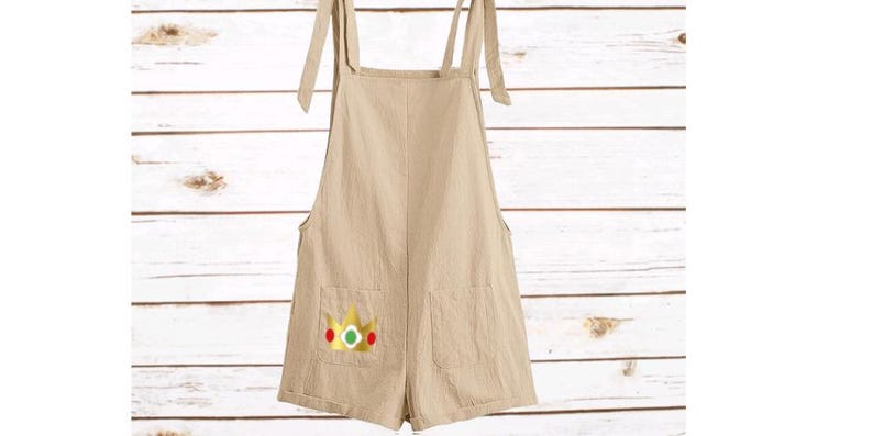 May include: A beige overall romper featuring tie-shoulder straps and two front pockets. The left pocket displays a graphic of a gold crown with a green center gem and red side accents, providing a distinct visual detail on the solid light-colored fabric.