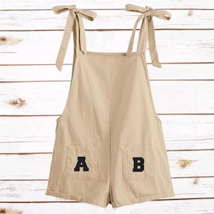 Op de afbeelding: Beige romper met strikbandjes en twee voorzakken. De zakken tonen de letters "A" en "B" in zwart. Gemaakt van een lichte stof, ideaal voor warm weer.