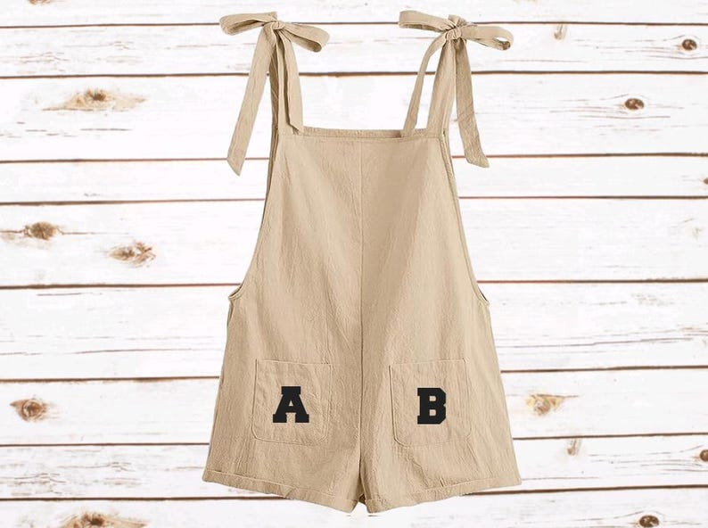 Op de afbeelding: Beige romper met strikbandjes en twee voorzakken. De zakken tonen de letters "A" en "B" in zwart. Gemaakt van een lichte stof, ideaal voor warm weer.