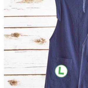 May include: A dark blue overall dress featuring shoulder straps that tie into bows, a square neckline, and a front pocket. A circular green patch with a white letter L is attached to the pocket. The garment has a straight hem with rolled cuffs.