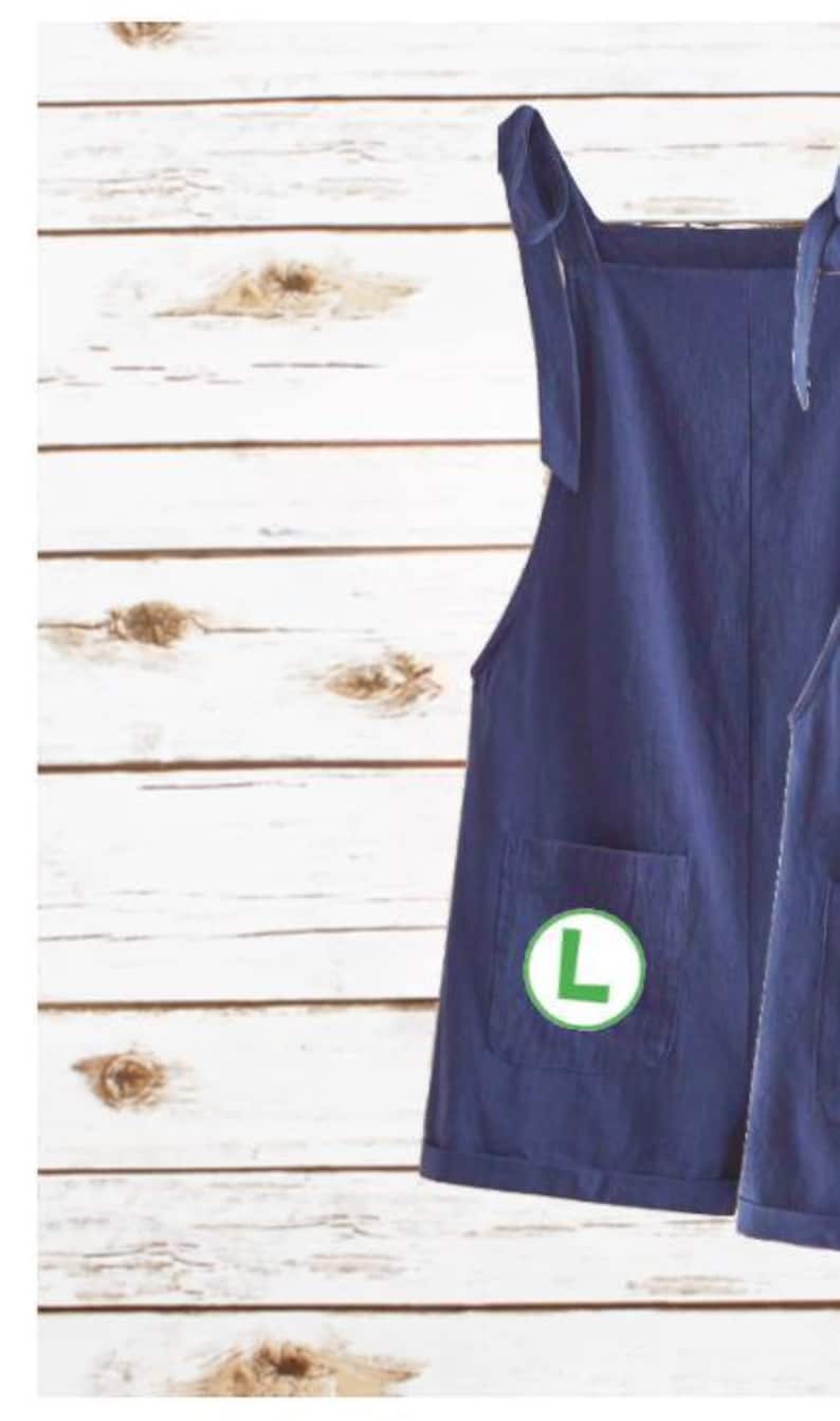 May include: A dark blue overall dress featuring shoulder straps that tie into bows, a square neckline, and a front pocket. A circular green patch with a white letter L is attached to the pocket. The garment has a straight hem with rolled cuffs.