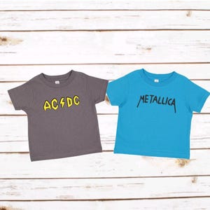 May include: A charcoal gray t-shirt with yellow AC/DC text and a lightning bolt graphic, alongside a bright blue t-shirt with black Metallica text. Both shirts feature short sleeves, a crew neckline, and a simple, straight-cut design.