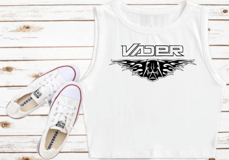 K&ouml;nnte beinhalten: Wei&szlig;es &auml;rmelloses Crop-Top mit einem schwarzen Grafikdruck von Darth Vaders Helm mit Flammen und dem Wort "VADER" dar&uuml;ber.