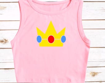 May include: A light pink sleeveless crop top featuring a centered graphic of a yellow crown with three blue oval accents and one central red oval accent. The garment has a round neckline and a straight hemline, showcasing a simple sleeveless design.