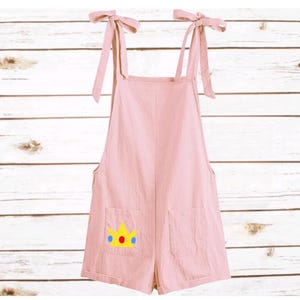 May include: A light pink romper featuring tie-shoulder straps and two front pockets. One pocket displays a yellow crown graphic with a red circle and two blue circles. The garment has a straight neckline and a short, loose-fitting silhouette.