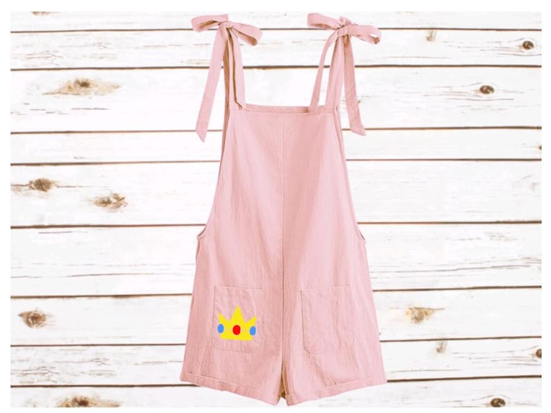May include: A light pink romper featuring tie-shoulder straps and two front pockets. One pocket displays a yellow crown graphic with a red circle and two blue circles. The garment has a straight neckline and a short, loose-fitting silhouette.