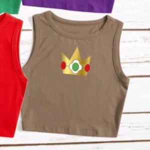 May include: A brown crop top with a gold crown graphic featuring red and green accents. The sleeveless top has a round neckline and is made of a soft material. Other colored tops are visible in the background.