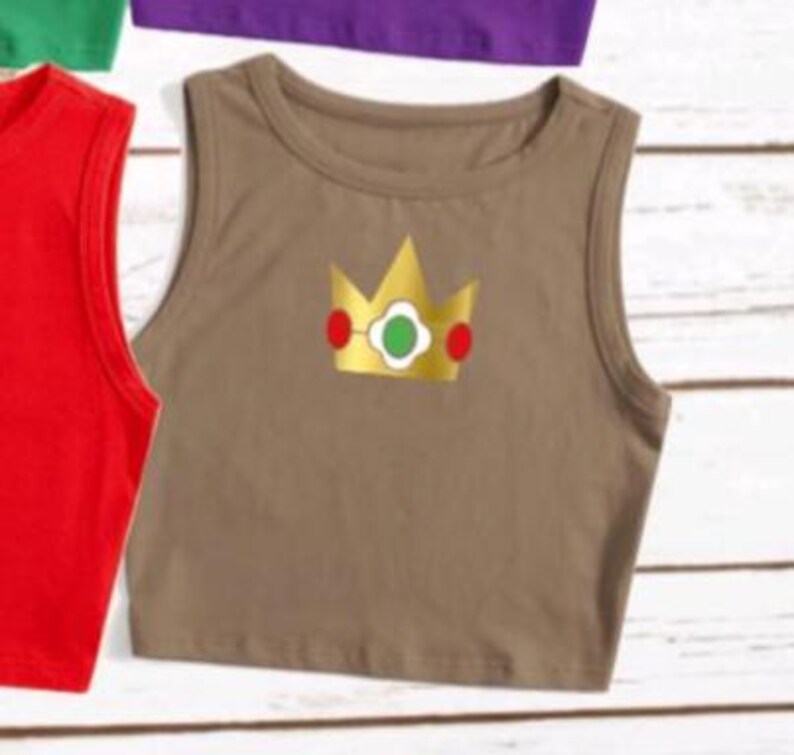 May include: A brown crop top with a gold crown graphic featuring red and green accents. The sleeveless top has a round neckline and is made of a soft material. Other colored tops are visible in the background.