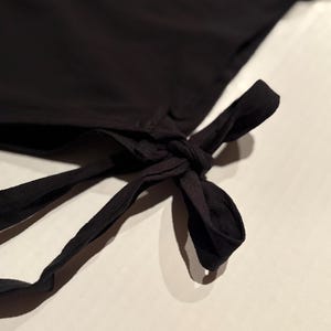 Op de afbeelding: Close-up van een zwart kledingstuk met een strikdetail. De stof lijkt een zacht, vloeiend materiaal te zijn. De strik is geknoopt en de uiteinden van de banden zijn zichtbaar. De achtergrond is licht en neutraal.